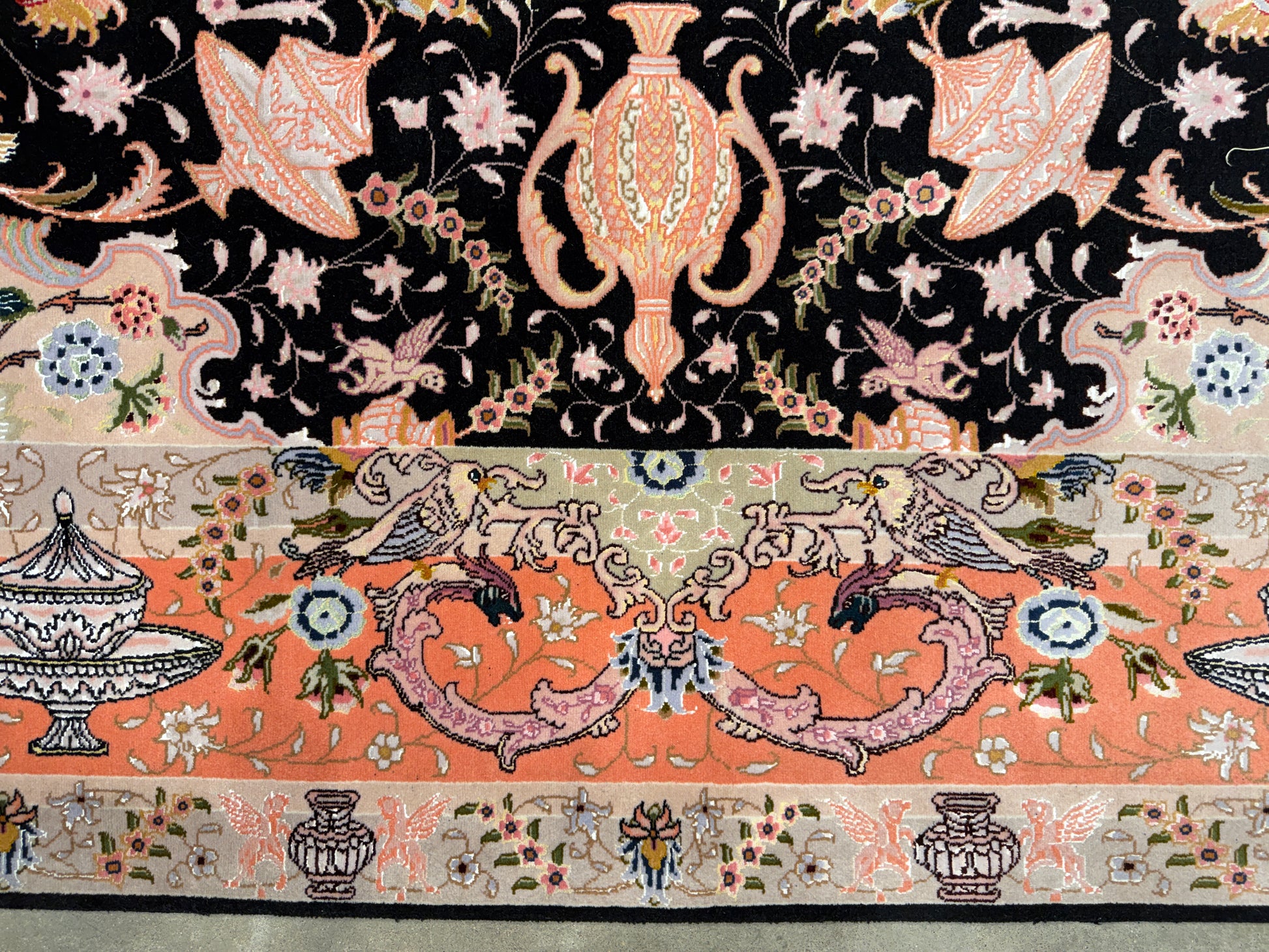 Close-up of an ornate patterned textile or rug showing a black upper field with peach floral motifs and a peach lower band with birds, urns, and floral details.