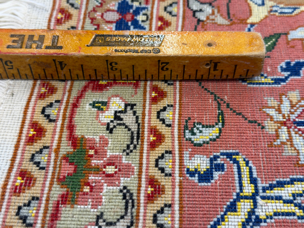 A close-up of a patterned rug with a wooden ruler laid across it for scale. The rug has a pink, beige, and blue floral and geometric design.