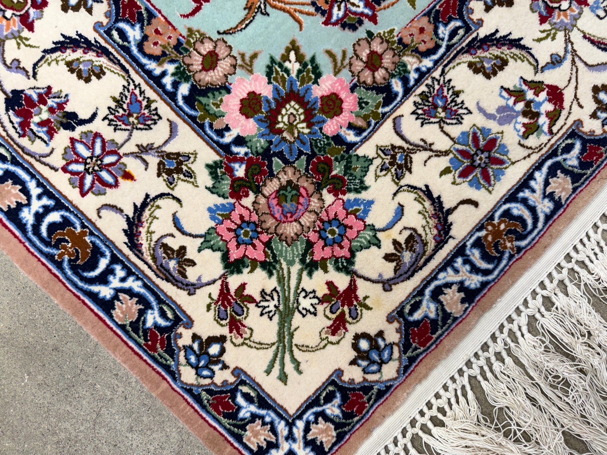 A close-up of an ornate Persian-style area rug corner, showing a cream field with a central floral motif in pink, blue, green, and red, surrounded by a detailed navy and burgundy border. White fringe is visible on the right side, laid over a gray floor.