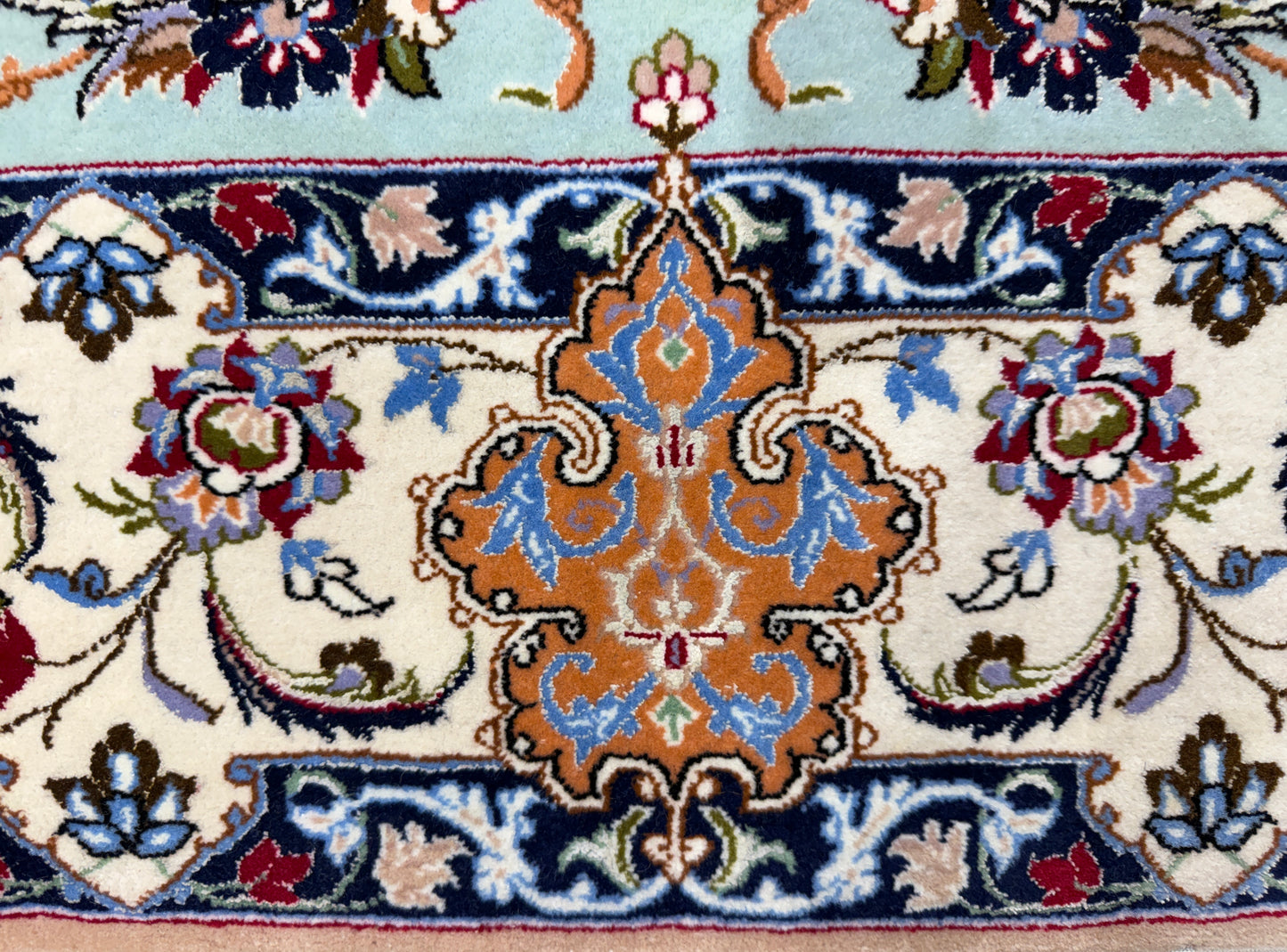 A close-up of a traditional patterned rug featuring a central ornate medallion in orange, blue, and cream, surrounded by floral motifs and bordered by navy and light blue sections.
