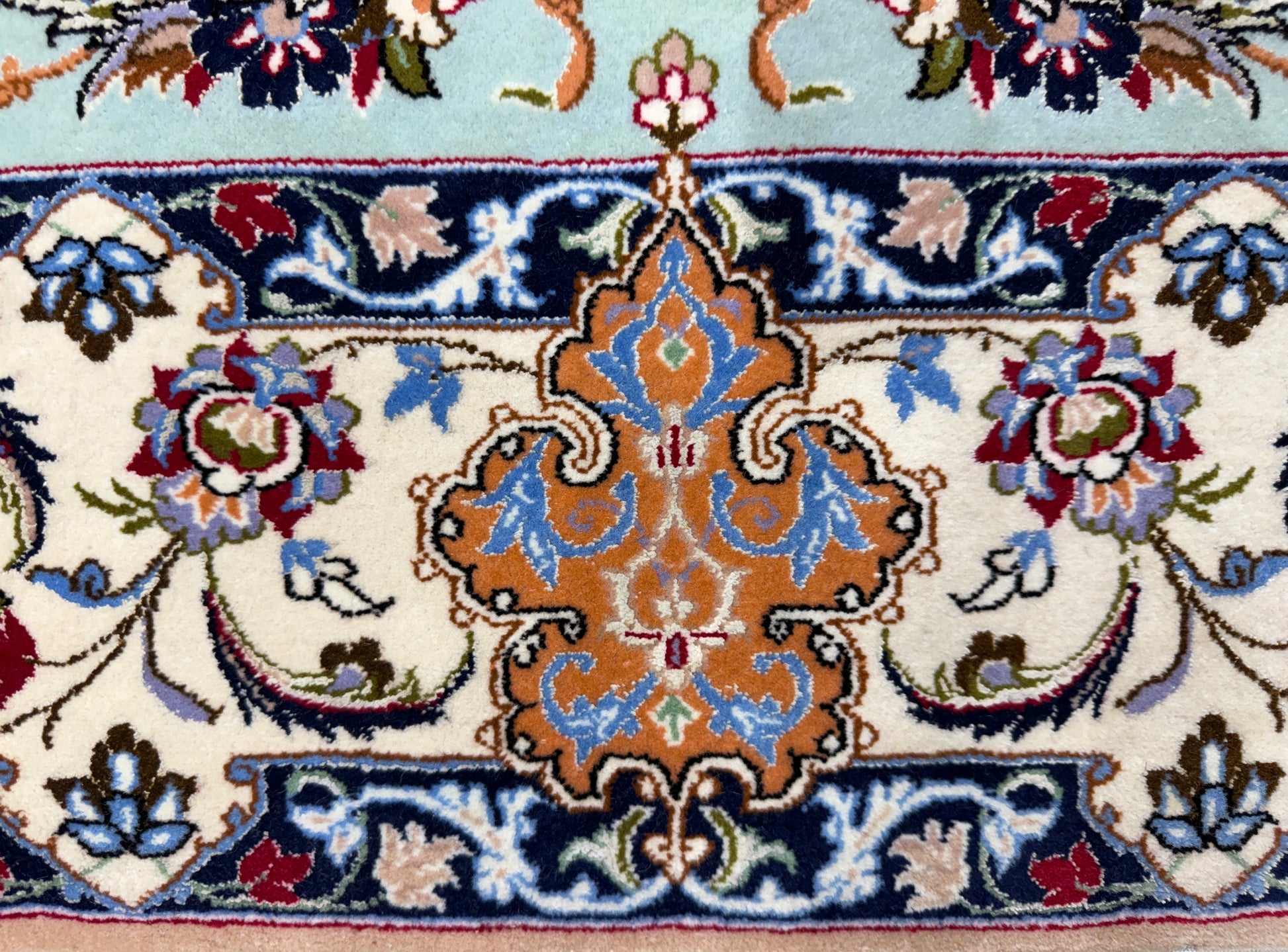 A close-up of a traditional patterned rug featuring a central ornate medallion in orange, blue, and cream, surrounded by floral motifs and bordered by navy and light blue sections.