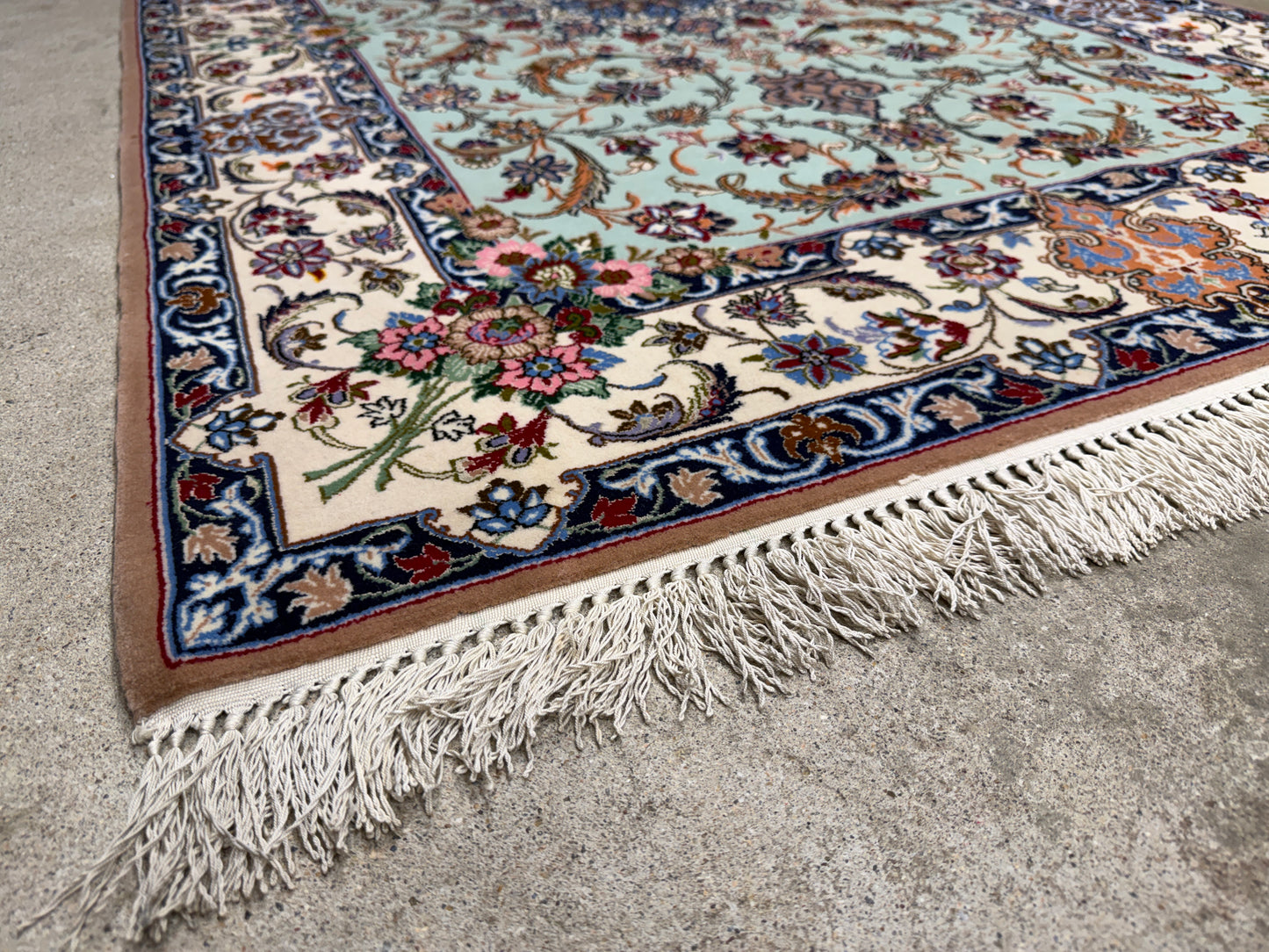 A close-up corner view of an ornate Persian-style area rug with cream, navy, blue, pink, and green floral motifs, resting on a gray concrete floor.