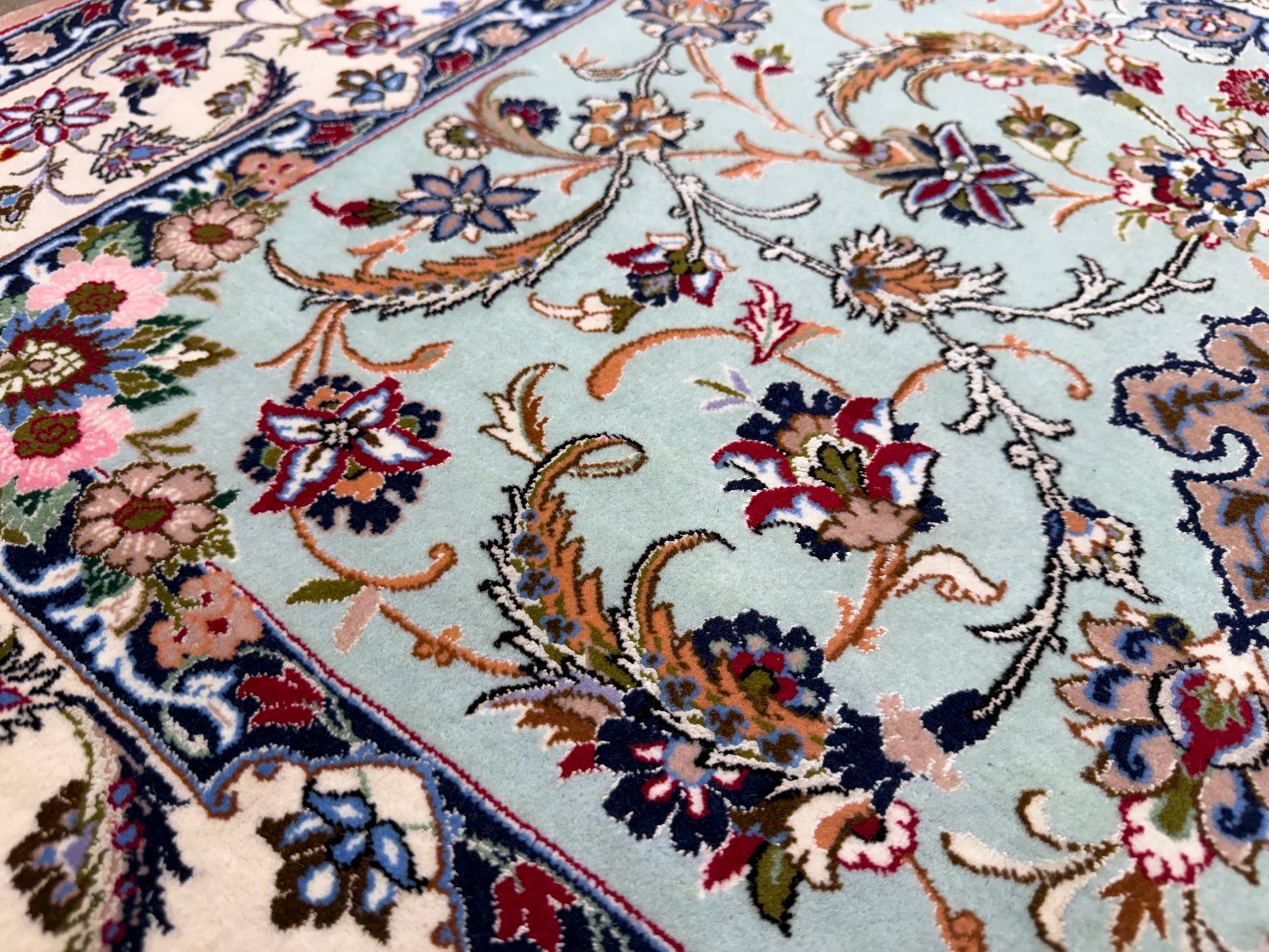 A close-up of an ornate Persian-style area rug with a light blue field, cream border, and intricate floral motifs in red, pink, green, and navy.